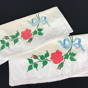 Puede incluir: Dos fundas de almohada de tela blanca con rosas rojas y hojas verdes bordadas. Cada funda de almohada tiene un lazo de cinta azul con ribete blanco. Las fundas de almohada tienen un ribete de encaje blanco alrededor de los bordes.