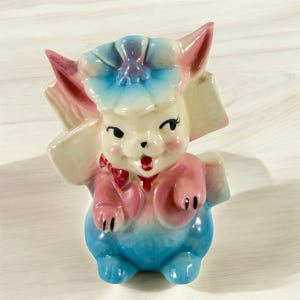 Könnte beinhalten: Vintage Keramikfigur eines lächelnden Tieres mit einem blütenförmigen Hut. Die Figur ist in Pastellfarben bemalt, darunter Rosa, Blau und Creme. Sie hat große Ohren und eine kleine rote Schleife. Die Figur ist ein Sammlerstück.