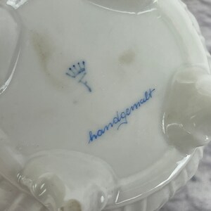 May include: A white porcelain dish with a blue crown and the words "handgemalt" in blue script.