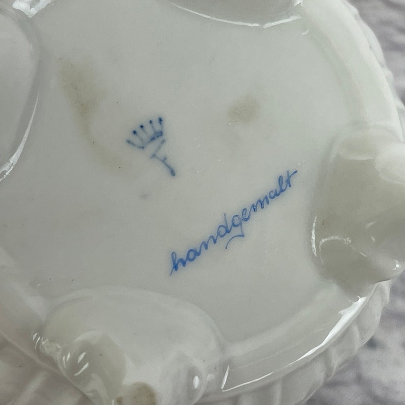May include: A white porcelain dish with a blue crown and the words "handgemalt" in blue script.