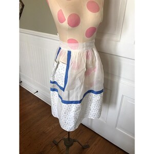 May include: A vintage white apron with blue trim and a pocket. The apron features a delicate pattern of small blue diamonds and is displayed on a mannequin with pink polka dots.