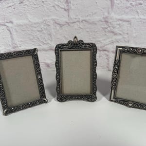Pode incluir: Três molduras retangulares feitas de metal em tom prateado escuro com padrões em relevo intrincados e pequenas pedras transparentes incrustadas. Cada moldura apresenta um design de borda decorativo distinto e uma abertura central.