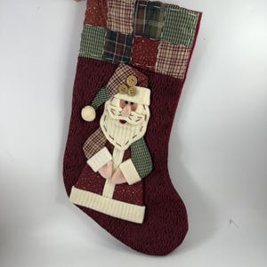 May include: Burgundy Christmas stocking with a Santa Claus applique. The stocking features a textured fabric and a patchwork cuff with green, red, and tan plaid squares. Santa has a white beard, a pink nose, and a green and tan hat.