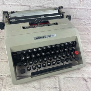May include: A white Olivetti Studio 45 typewriter with a black keyboard and red tab key. The typewriter has a black carriage return lever and a black paper release lever.