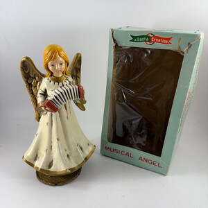 May include: A vintage musical angel figurine with blonde hair, wearing a white dress with gold accents and holding a red and black accordion. The angel is standing on a gold base and is in its original box, which is blue and white with the text "A Santa Creation. Musical Angel".