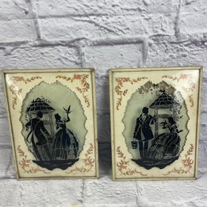 Può includere: Due opere d'arte vintage con silhouette incorniciate. Ognuna presenta una silhouette nera di una coppia vicino a un pozzo, con accenti floreali e di foglie. Le cornici sono bianco sporco con un aspetto invecchiato.