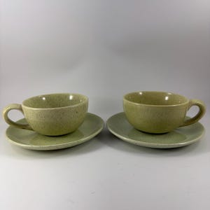 Puede incluir: Dos tazas de té y platillos de cerámica verde oliva moteado. Las tazas tienen forma redondeada con asa. Los platillos son redondos y planos. El juego se exhibe sobre un fondo blanco.