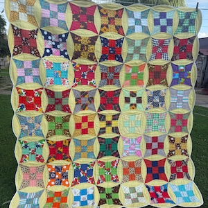 May include: A handmade patchwork quilt featuring a grid of multi-colored fabric squares in various patterns, including floral, plaid, and geometric prints. Each square is framed by curved, pale yellow fabric borders, creating a scalloped edge.