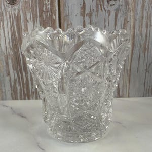May include: A clear glass vase with a scalloped rim and intricate cut glass design. The vase has a starburst pattern and is perfect for displaying flowers.