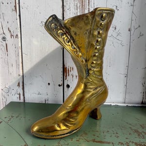 Pode incluir: Um item decorativo vintage em forma de bota de latão. A bota apresenta um salto alto, detalhes de botões e padrões em relevo intrincados. O latão tem um tom dourado quente, e a bota está colocada sobre uma superfície verde.