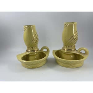 May include: A pair of pale yellow ceramic candle holders featuring a rounded base with a woven-style texture, a small side handle, and a central pillar with gold-toned accents. The candle holders have a smooth, glossy finish and a uniform design.