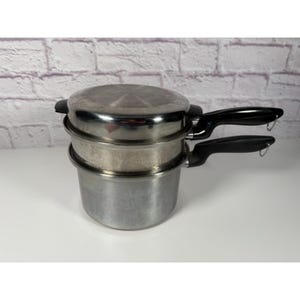 Puede incluir: Un juego de ollas vaporeras de acero inoxidable con tapa y dos asas negras. El juego incluye una olla base, un inserto para vapor y una tapa. La olla está diseñada para cocer al vapor verduras, mariscos u otros alimentos.