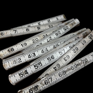 May include: A collection of white folding rulers with black markings, displaying inch measurements. The rulers are arranged at various angles against a black background, showing numbers from 1 to 70.