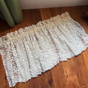 May include: Cream-colored lace curtain panel. The curtain is gathered at the top and features a delicate floral pattern. The lace is semi-sheer, allowing light to filter through. The curtain is lying on a wooden surface.