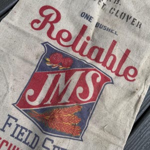 May include: A vintage beige canvas bag with the word "Reliable" in red script. The bag features a red, white, and blue emblem with "JMS" and illustrations of wheat and berries. Text includes "One Bushel" and "Field Seeds".