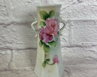 May include: A white porcelain vase with two handles and a floral design. The vase is decorated with pink and green roses.