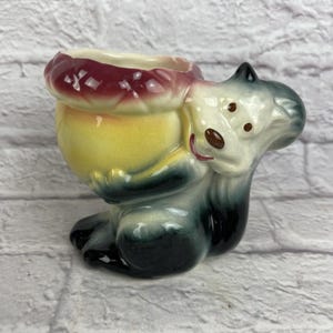 Puede incluir: Figura vintage de cerámica de ardilla, sosteniendo un recipiente amarillo y rojo. La ardilla está pintada en tonos de gris, blanco y negro, con detalles en marrón. El recipiente tiene un borde con volantes. Decoración.