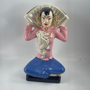Puede incluir: Figura de cerámica de una mujer con chaqueta rosa con diseño de dragón blanco, pantalón azul y abanico. La figura tiene cabello negro, labios rojos y detalles dorados. La base es negra con ribete dorado.