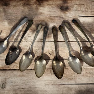 Op de afbeelding: Een bovenaanzicht van zeven antieke zilveren lepels, gerangschikt in een halve cirkel op een verweerd houten oppervlak. De lepels variëren in patina, met enkele die aanzienlijke slijtage en verkleuring vertonen.