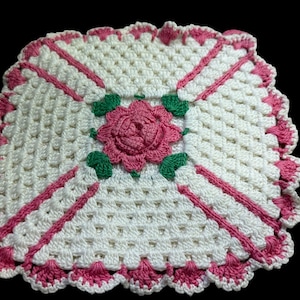May include: A square, handmade crochet doily featuring a central pink rose with green leaves. The doily has a white background with pink accents and a scalloped edge, showcasing a vintage aesthetic.