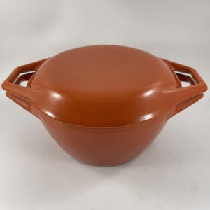 May include: A round, orange-brown casserole dish with a matching lid. The dish has two rectangular handles on either side. The lid fits snugly on top, creating a smooth, seamless appearance. The dish is made of a durable material.