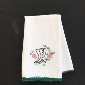May include: White hand towel with a green crocheted edge. The towel features an embroidered design of a black boot with a floral arrangement in pink, blue, and purple. The towel is folded in half.