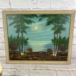 May include: An original oil painting of a moonlit lake scene. The artwork features tall trees on either side of the lake, with a full moon in the sky. The painting is framed in a light-colored wooden frame.