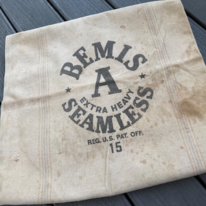 May include: Vintage beige fabric with the text "BEMIS A EXTRA HEAVY SEAMLESS" in black. The fabric has stains and a striped pattern. The number "15" is printed below the text. The fabric is likely from a bag or sack.