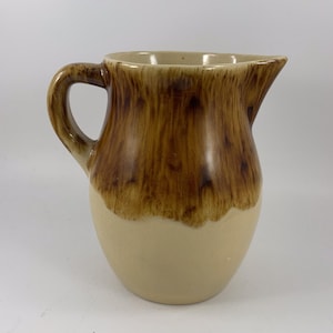 May include: A vintage ceramic pitcher with a handle and spout. The top half is a rich brown color with a textured, streaked appearance, while the bottom half is a creamy beige. The pitcher has a classic, rounded shape.