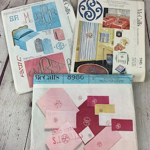 May include: Three vintage McCall's transfer patterns for embroidery. The patterns feature a script alphabet, a monogram design, and a single scroll design. The patterns are printed on white paper with blue and red accents.