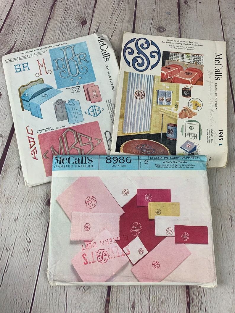 May include: Three vintage McCall's transfer patterns for embroidery. The patterns feature a script alphabet, a monogram design, and a single scroll design. The patterns are printed on white paper with blue and red accents.