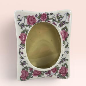 Könnte beinhalten: Ein weißer Keramik-Bilderrahmen mit floralem Design. Der Rahmen hat eine ovale Öffnung und ist mit rosa Rosen, grünen Blättern und kleinen lila Blumen verziert. Der Rahmen hat eine leicht geschwungene Form.