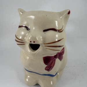 Peut inclure: Figurine de chat vintage en céramique avec un visage souriant, des moustaches brunes et un nœud papillon bordeaux. Le chat a un collier bleu et une petite ouverture pour la bouche. La figurine est blanc cassé avec des accents bruns et bordeaux.
