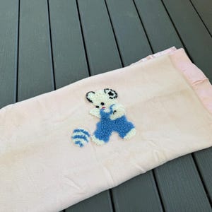 May include: A light pink baby blanket with a satin edge. The blanket features an embroidered design of a white teddy bear wearing blue overalls and playing with a blue and white striped ball. The blanket is folded.