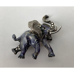 May include: A vintage elephant brooch with a silver-tone metal head, trunk, and tusks. The elephant's body is a mottled blue with darker blue and brown accents. The brooch has a pin closure on the back.