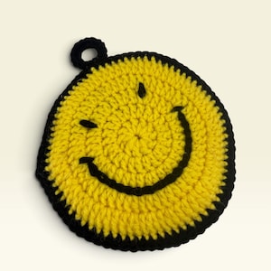 May include: A circular yellow crochet coaster featuring a black stitched smiley face design with two small eyes and a wide curved mouth. The coaster is edged with a thick black border and includes a small black circular loop at the top for hanging.