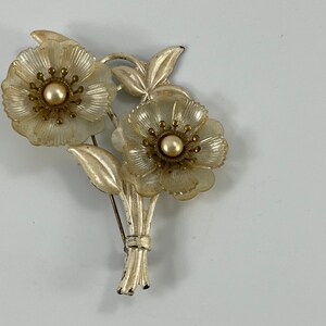 May include: A vintage brooch featuring two white plastic flowers with gold centres and pearl accents. The flowers are attached to a gold-toned stem with leaves.