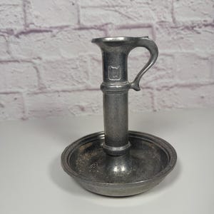 Peut inclure: Un bougeoir en étain vintage avec une poignée. Le bougeoir a une base ronde et une tige cylindrique haute avec une poignée. Le métal a un aspect vieilli et terni.