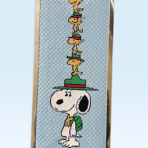Peut inclure: Une pièce d'art en point de croix encadrée rectangulaire représentant un beagle blanc avec une oreille noire et un sac à dos vert. Quatre oiseaux jaunes, portant chacun un chapeau vert à bande rouge, sont empilés sur la tête du beagle sur fond bleu.