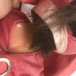 May include: Close-up of a pink fabric doll with brown hair. The doll is wearing a pink outfit with a white trim. The doll's neck has a small, faded label with the text "Made in Hong Kong".