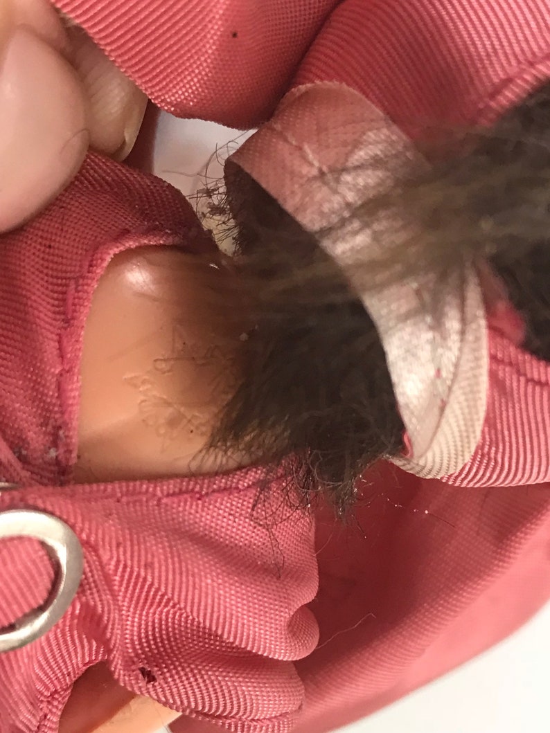 May include: Close-up of a pink fabric doll with brown hair. The doll is wearing a pink outfit with a white trim. The doll's neck has a small, faded label with the text "Made in Hong Kong".