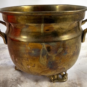 May include: A round brass planter with a polished rim, two curved side handles, and three ornate feet. The metal surface features a weathered, multi-toned patina with visible dark oxidation spots and warm golden highlights throughout the body.