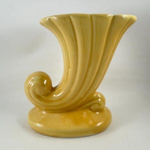 May include: A yellow ceramic vase with a curved, horn-like shape. The vase features a flared top and a base with a rounded, scroll-like design. The vase is approximately 8 inches tall.