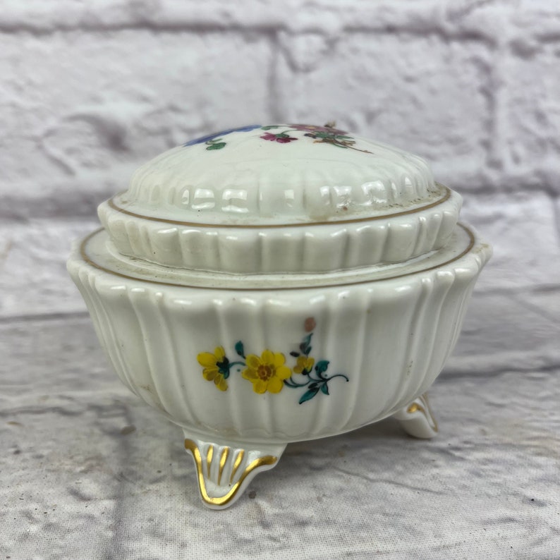 May include: A white porcelain trinket box with a floral design and gold accents. The box has a lid and three feet.