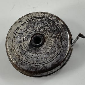 Puede incluir: Un antiguo metro de cinta circular de metal. El texto "PAL'S MECHANICS" y "6 FEET" están grabados en la superficie. El metro de cinta es de color plateado deslustrado con una pequeña lengüeta metálica que se extiende desde el lateral.