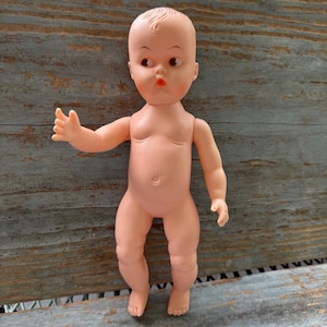Könnte beinhalten: Eine Vintage-Baby-Puppe aus Kunststoff in Pfirsichfarbe. Die Puppe hat ein detailliertes Gesicht mit bemalten Augen, rosigen Wangen und einem kleinen Mund. Die Arme der Puppe sind ausgestreckt, und sie steht auf einer Holzoberfläche.
