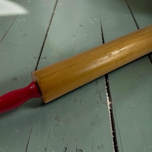 May include: A wooden rolling pin featuring a smooth, natural light brown cylindrical barrel and two bright red painted handles on each end. The rolling pin has a classic design with tapered handles and a central rotating shaft for dough preparation.