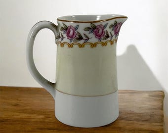 May include: A small white porcelain creamer with a handle. The creamer has a floral design with pink roses and green leaves around the top edge. The design is accented with gold trim.