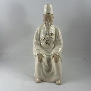 以下が含まれることがあります： 座った姿勢の、年老いたアジア人男性の陶器製フィギュア。 金色のトリムと高い帽子が付いた白いローブを着ています。 フィギュアは長い白いひげがあり、金色のアクセントで装飾されています。