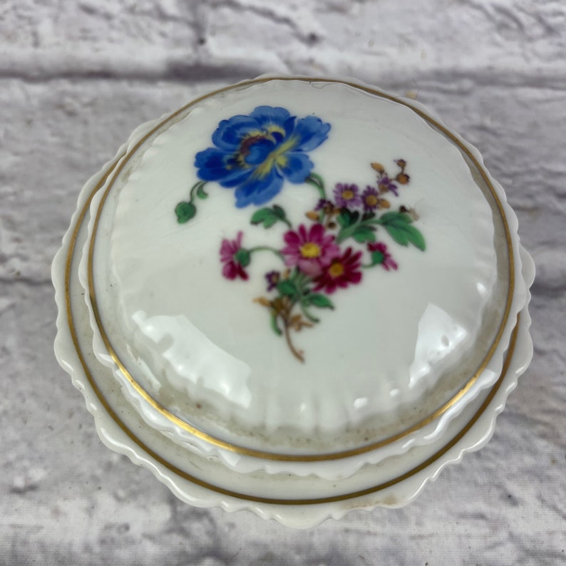 May include: White porcelain trinket box with a gold trim and a floral design. The lid features a blue flower with pink and white flowers surrounding it.